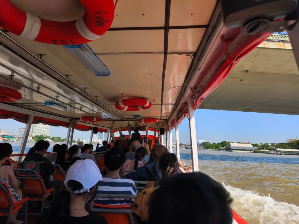 Vista desde el interior de un barco del transporte fluvial en Bangkok, con pasajeros sentados a ambos lados de la embarcación, el río Chao Phraya extendiéndose al frente y la brisa del agua, mientras el barco navega entre muelles, una de las formas más auténticas de moverse por la ciudad.