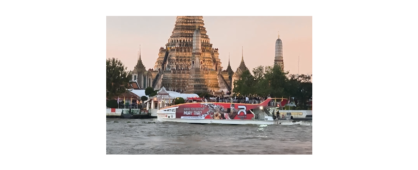 Vista desde lejos de un barco de bandera azul del transporte fluvial en Bangkok navegando por el río Chao Phraya frente a Wat Arun, el Templo del Amanecer, con sus agujas iluminándose al atardecer, una de las estampas más emblemáticas del río y un trayecto turístico de 40 baht.