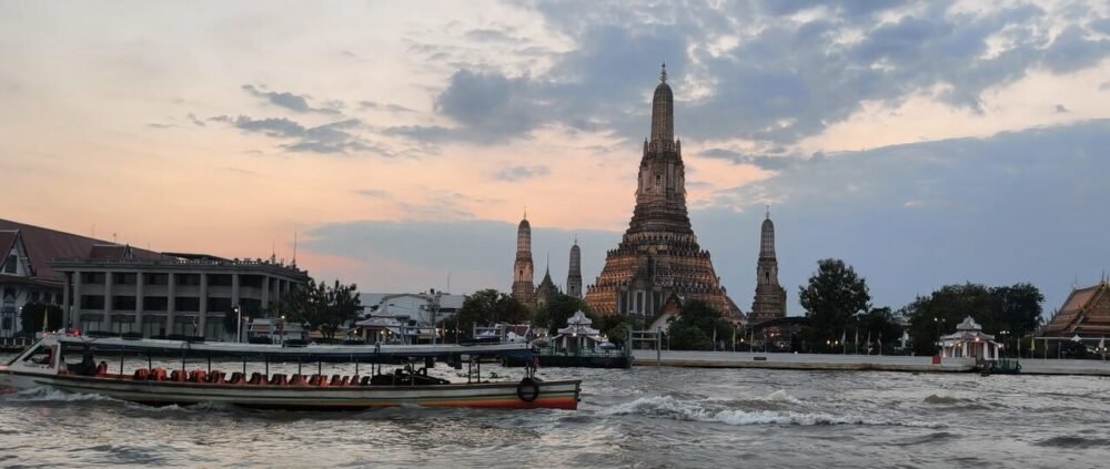 Barco de bandera naranja del transporte fluvial en Bangkok navegando por el río Chao Phraya frente al Wat Arun, el Templo del Amanecer, mientras sus agujas empiezan a iluminarse al atardecer, una de las estampas más mágicas de la ciudad y un trayecto de solo 16 baht.