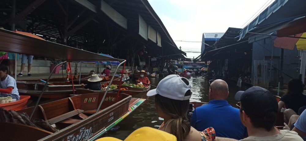 Vista de varias barcas de madera en el mercado flotante de Damnoen Saduak, uno de los destinos más emblemáticos del transporte fluvial en Bangkok y sus alrededores. En la imagen se ven tanto barcas de cola larga (motorizadas) como barcas de remo, cargadas de frutas, verduras y productos locales, mientras los vendedores y compradores negocian desde el agua, ofreciendo una experiencia única que combina comercio y tradición.